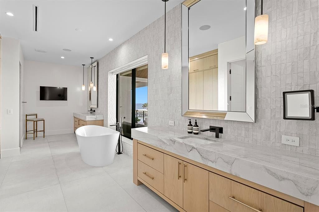 This is a luxurious primary bathroom featuring a modern freestanding bathtub, a spacious vanity with marble countertops and light wood cabinetry, and large mirrors with pendant lighting. The bathroom has a clean, contemporary design with white and gray tones, creating a serene and spa-like atmosphere. A sliding glass door leads to an outdoor balcony.