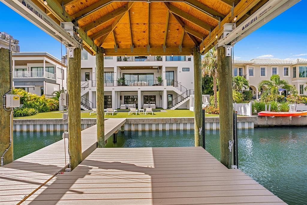 This image showcases the rear exterior of a luxurious waterfront property, viewed from a wooden dock. The multi-story home features multiple balconies, large windows, and manicured landscaping leading down to the water. The dock itself is constructed of light-colored wood and supported by sturdy pilings, creating a serene and inviting atmosphere.