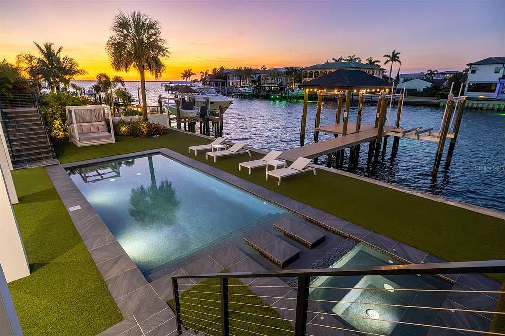 This image showcases a luxurious backyard featuring a modern pool and spa area with waterfront access. The pool is surrounded by sleek tiling and artificial turf, complemented by lounge chairs and a private dock with a gazebo. The scene is set against a vibrant sunset, enhancing the property's appeal and creating a sense of relaxation and upscale living.