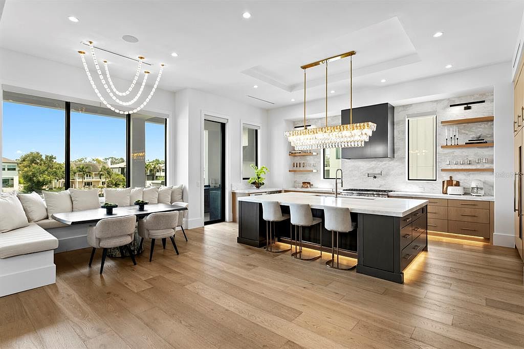 This is a bright and spacious kitchen and dining area featuring a large island with bar seating, modern cabinetry, and high-end appliances. The open floor plan connects the kitchen seamlessly with the dining area, which includes a built-in bench and a stylish chandelier. The hardwood flooring and ample natural light create a warm and inviting atmosphere.