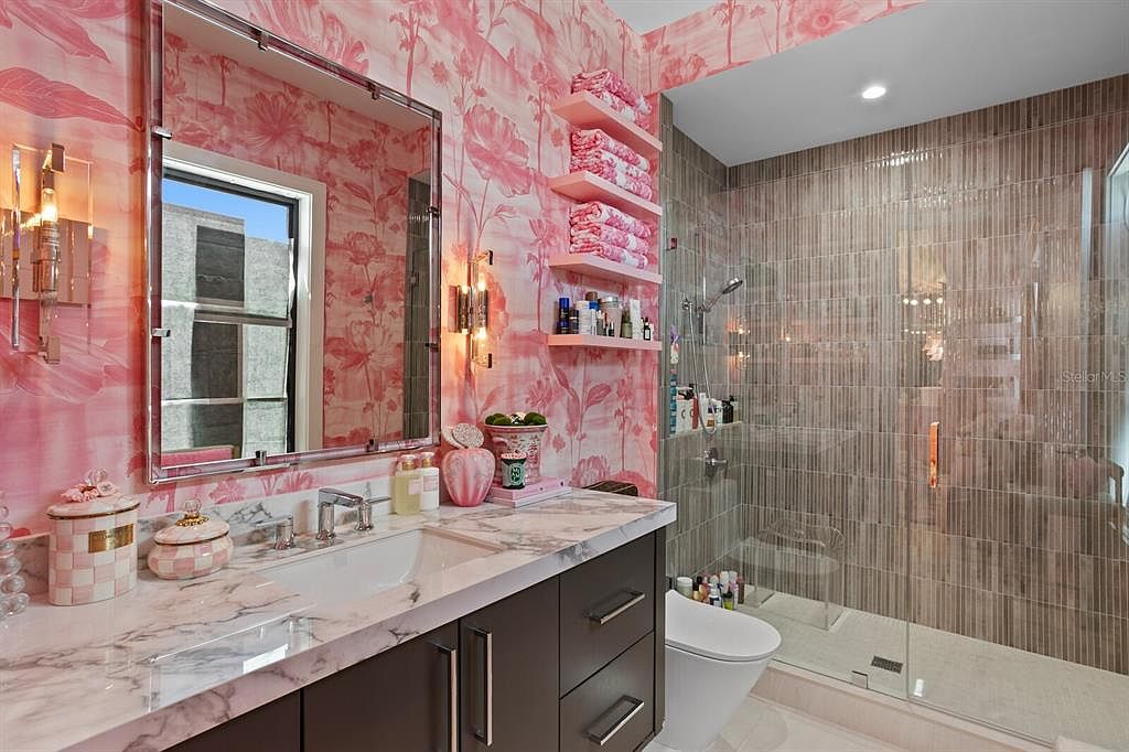 This is a stylish primary bathroom featuring pink floral wallpaper, a marble countertop vanity with dark brown cabinetry, and a glass-enclosed shower with vertically stacked tiles. Pink shelves hold neatly folded towels, complementing the overall color scheme. The design creates a luxurious and visually appealing space.