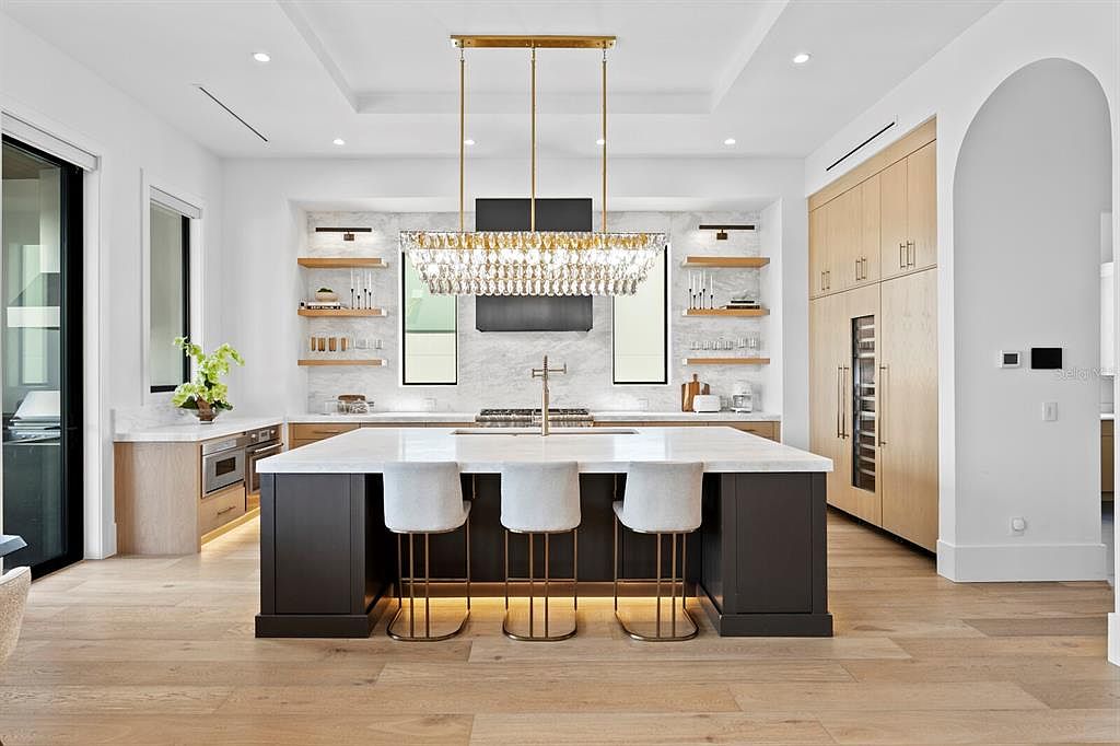 This is a bright and modern kitchen featuring a large island with seating, a statement chandelier, and custom cabinetry. The kitchen is designed with a clean aesthetic, incorporating light wood floors, white countertops, and stainless steel appliances. Open shelving and a marble backsplash add to the luxurious feel of the space.