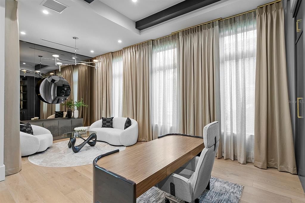 This is an interior shot of a modern office space featuring a large wooden desk with a gray office chair, complemented by two curved white sofas and a unique glass coffee table on a round rug. The room is well-lit with natural light filtering through large windows covered by elegant beige curtains, creating a sophisticated and comfortable work environment.