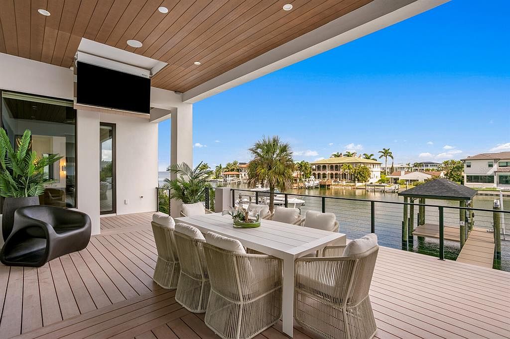 This image showcases a luxurious outdoor patio/deck area with a stunning waterfront view. The space is furnished with a modern white dining table and woven chairs, perfect for entertaining. A large flat-screen TV is mounted under the covered ceiling, and the deck is made of wood, adding warmth and texture to the scene. The overall impression is one of upscale outdoor living and relaxation.