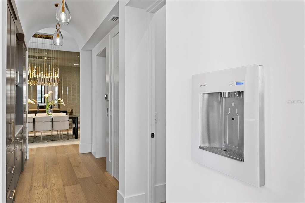 This interior shot showcases a modern hallway with light wood flooring and white walls. A built-in water dispenser is mounted on the wall, adding a unique feature. The hallway leads to a dining area with a chandelier, creating a sense of depth and luxury.