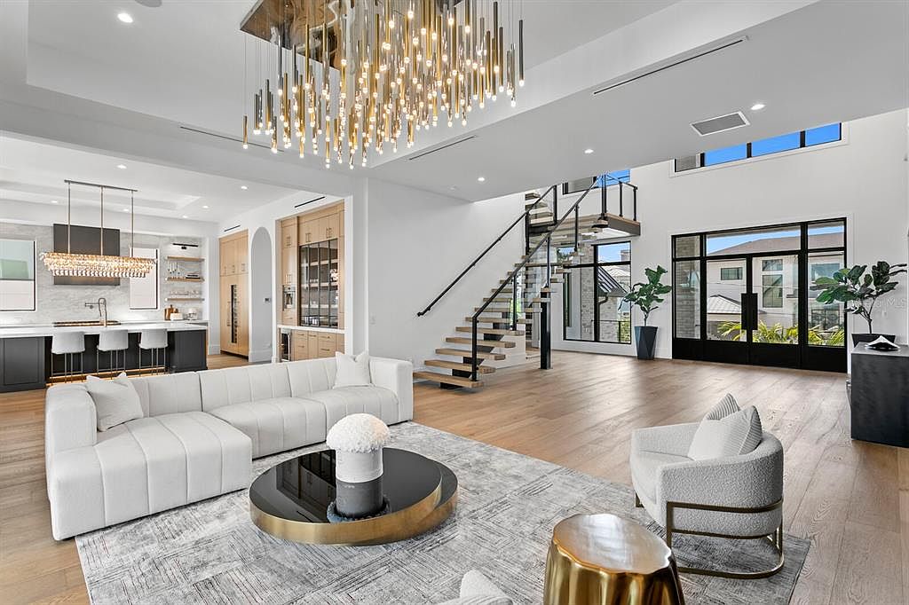 This is a spacious and luxurious living room featuring a large white sectional sofa, a modern chandelier, and a stylish coffee table. The open floor plan seamlessly connects to the kitchen and staircase, enhancing the sense of flow and connectivity. Large windows provide ample natural light, highlighting the hardwood floors and contemporary design elements.