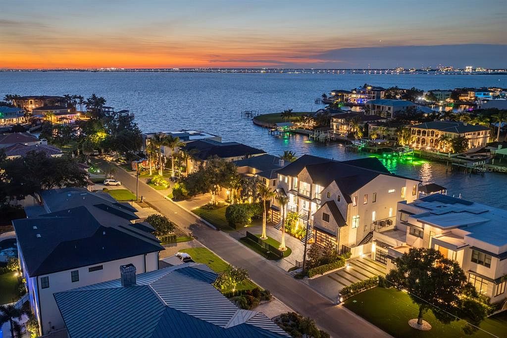 This aerial view showcases a luxurious waterfront neighborhood at dusk, featuring modern homes with manicured lawns and illuminated exteriors. The scene captures the tranquil water, a distant bridge, and a vibrant sunset, creating an impression of upscale coastal living. The perspective is from above, providing a comprehensive view of the neighborhood's layout and proximity to the water.