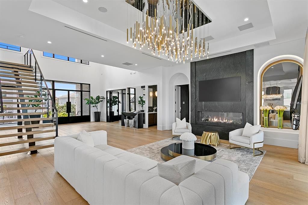 This is a spacious and luxurious living room featuring a modern design. The room is bright with natural light and features a large white sofa, a fireplace with a black stone surround, and a unique chandelier. The open floor plan and hardwood floors contribute to the room's elegant and inviting atmosphere.