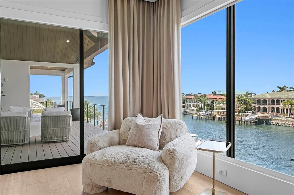 This interior shot showcases a luxurious living room with a plush, oversized armchair positioned near a large window offering a stunning waterfront view. The room features neutral tones, with elegant curtains and a small side table adding to the sophisticated ambiance. The open layout extends to an outdoor deck visible through a sliding glass door, enhancing the sense of spaciousness and connection to the surrounding environment.