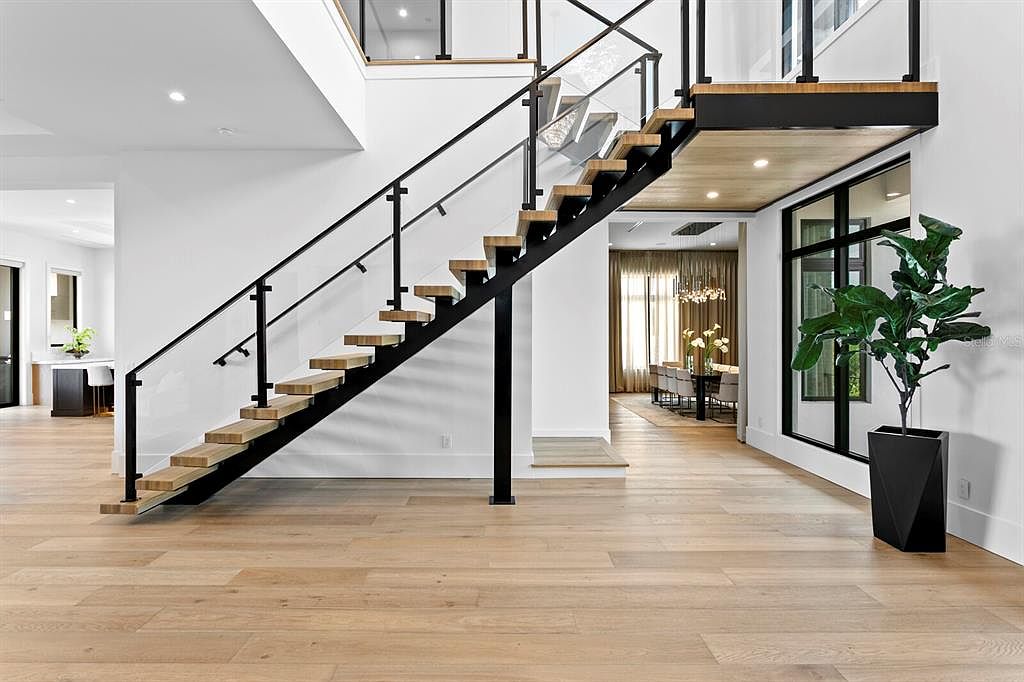 This interior shot showcases a modern hallway with a striking staircase featuring wooden steps and black metal railings with glass panels. The light hardwood flooring adds warmth to the space, while a large potted plant near the entrance brings a touch of nature indoors. The overall impression is clean, contemporary, and inviting.
