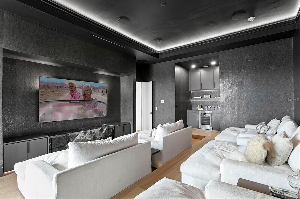 This is a modern living room featuring a large television mounted on a textured gray wall, complemented by gray cabinets and a marble-like accent piece. The room includes two white sofas and a built-in bar area with gray cabinets and a wine fridge. The overall aesthetic is sleek and luxurious, with recessed lighting enhancing the ambiance.