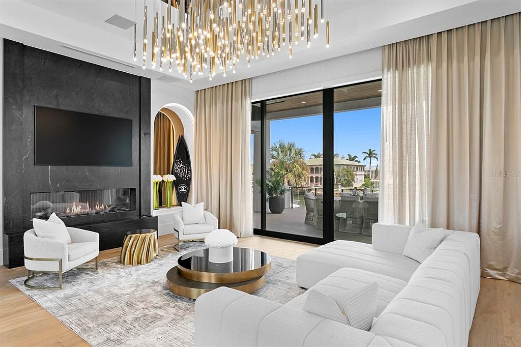 This is a luxurious living room featuring a modern fireplace with a large flat-screen TV above it. The room is decorated in neutral tones with white furniture, a unique chandelier, and large windows that offer a view of the outdoors. The overall impression is one of elegance and sophistication.