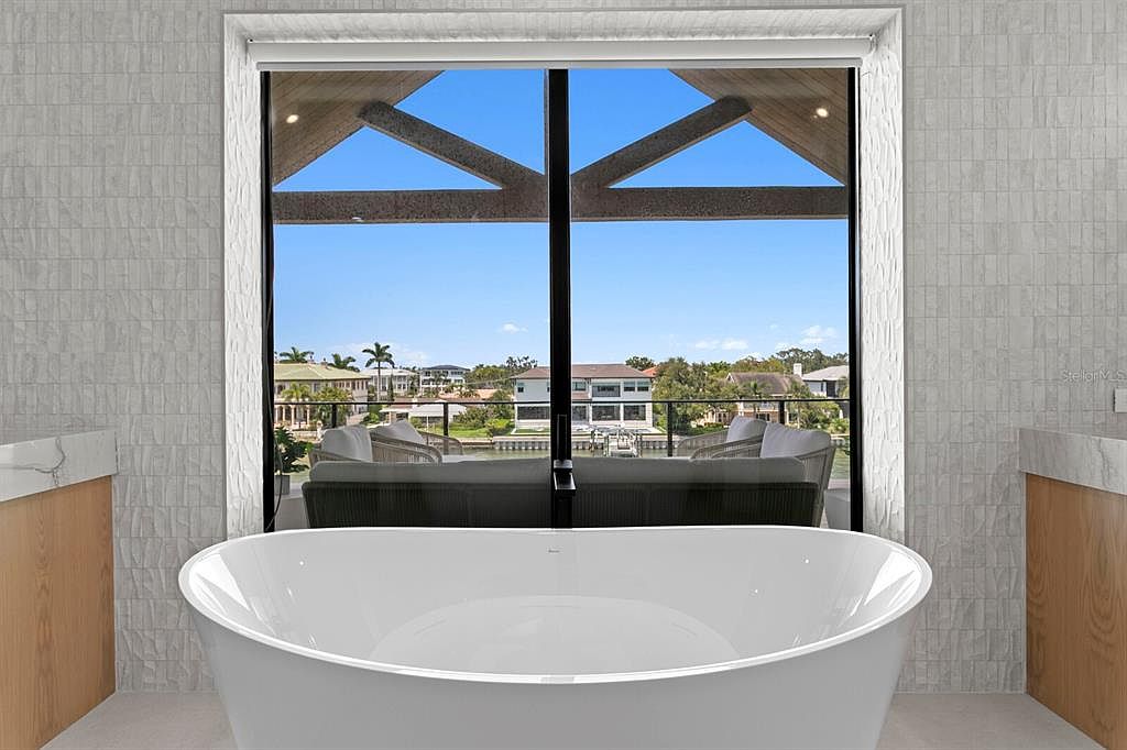 This is a luxurious primary bathroom featuring a freestanding white bathtub positioned in front of a large window with a view of waterfront homes. The bathroom has light gray textured walls and light wood vanities with white countertops on either side of the tub. The scene is designed to highlight the relaxing atmosphere and the beautiful view.