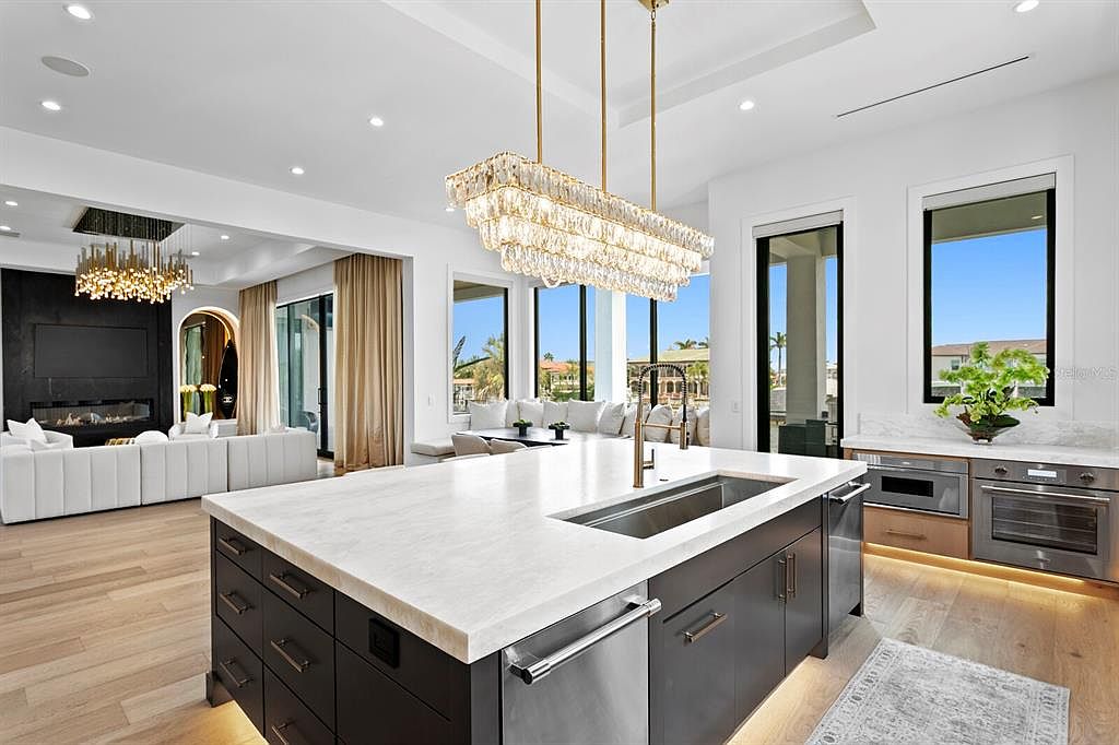 This is a bright and modern kitchen featuring a large island with a white countertop and dark cabinetry. The kitchen is open to the living area, with views of the water through large windows. A statement chandelier hangs above the island, adding a touch of elegance.