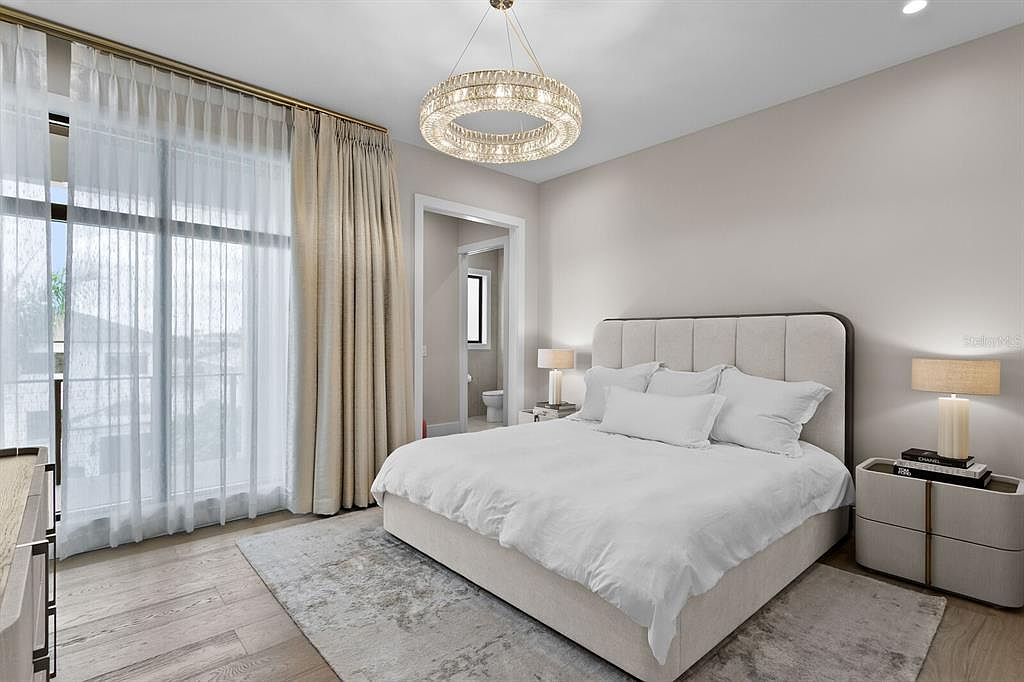 This is a bright and airy primary bedroom featuring a large bed with white linens, a neutral color palette, and a modern chandelier. The room has large windows with sheer and blackout curtains, providing ample natural light and privacy. The overall impression is one of comfort and sophistication.