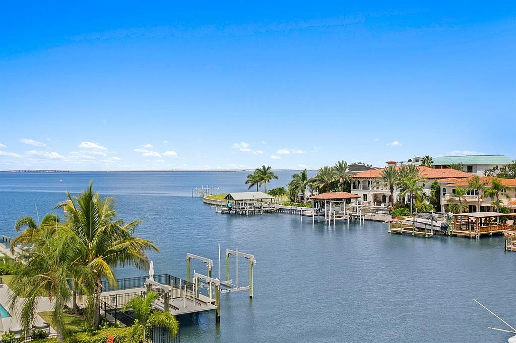 This aerial view showcases a luxurious waterfront property with Mediterranean-style architecture, featuring red-tiled roofs and multiple balconies. The property includes private docks with boat lifts, surrounded by calm, blue waters. Lush palm trees and manicured landscaping enhance the tropical ambiance, creating an exclusive and serene setting.