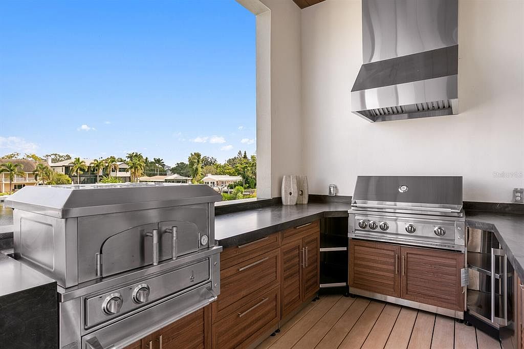 This image showcases an outdoor kitchen on a patio or balcony, featuring stainless steel appliances including a pizza oven and a grill. The kitchen is built with wooden cabinets and dark countertops, offering a modern and functional space for outdoor cooking and entertaining. The view from the patio includes a waterfront scene with houses and trees in the background.