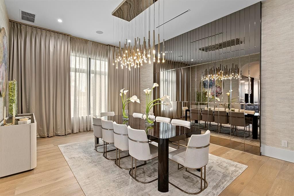 This is an elegant dining room featuring a long, dark wood table with seating for eight, complemented by modern chairs with light upholstery and gold accents. A striking chandelier hangs above the table, and a large mirrored wall visually expands the space. The room is bathed in soft, natural light filtering through sheer curtains, creating a sophisticated and inviting atmosphere.
