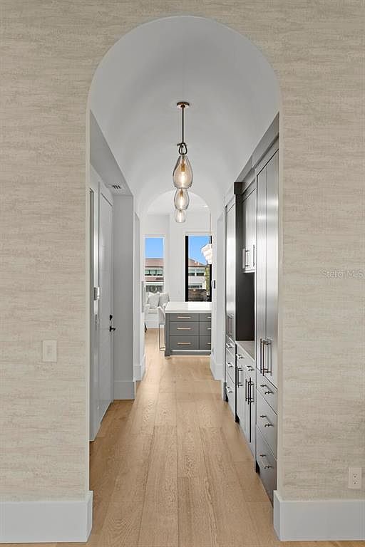 This interior shot showcases a hallway with light wood flooring and textured wallpaper. An arched entryway frames the view into a bright space, while modern pendant lighting adds a touch of elegance. Built-in cabinetry lines one side of the hallway, providing ample storage.