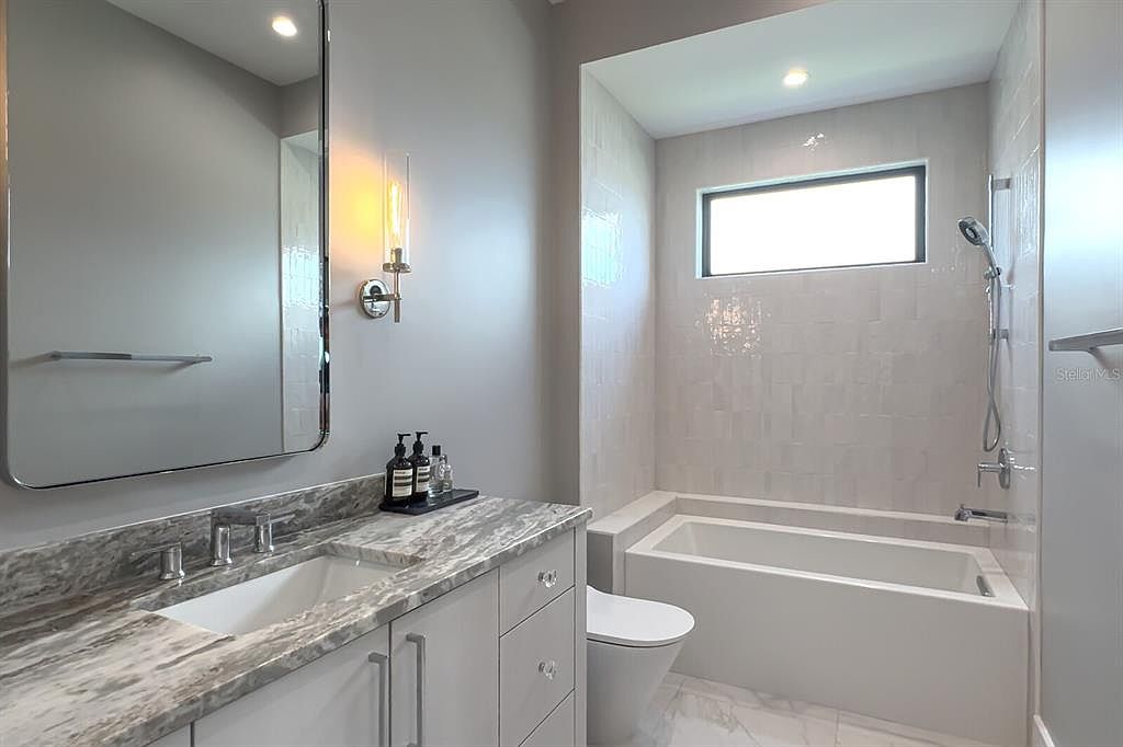 This is a modern bathroom featuring a gray and white color scheme. The vanity has a marble countertop and white cabinets with crystal knobs. A large mirror hangs above the sink, and a bathtub is located next to the toilet, with a window above the tub providing natural light.