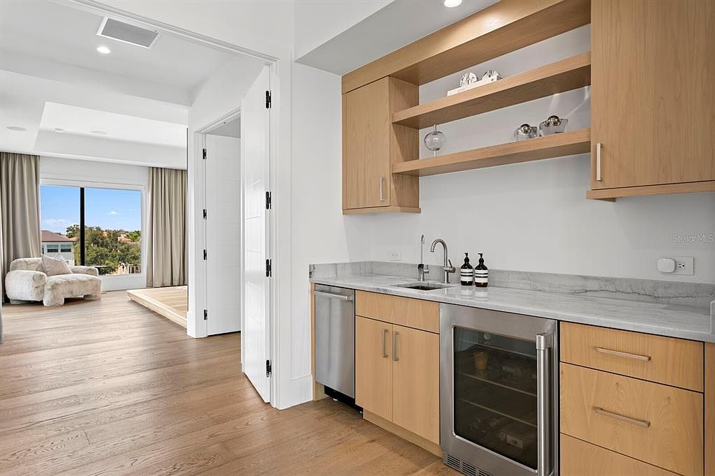 This interior shot showcases a modern living space with a built-in bar area featuring light wood cabinetry, open shelving, and a wine refrigerator. The room extends into a seating area with a plush armchair and a view through a large window, creating a bright and inviting atmosphere. The hardwood flooring adds warmth and continuity to the space.