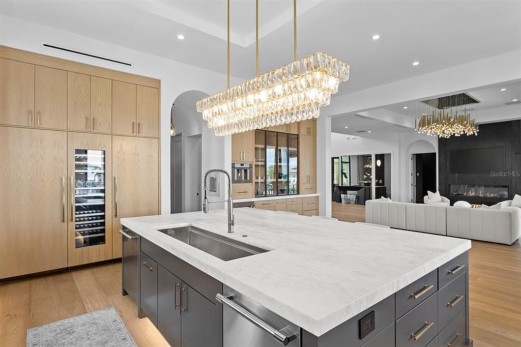 This is a bright and modern kitchen featuring a large island with a white countertop and gray cabinetry. The kitchen includes stainless steel appliances, a wine cooler, and custom wood cabinetry. A rectangular crystal chandelier hangs above the island, and the open floor plan leads to a living area with a fireplace.