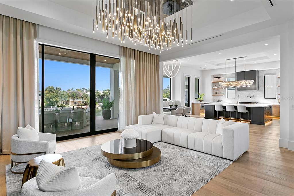 This is a luxurious living room featuring a large white sectional sofa, a round coffee table with gold accents, and two armchairs. The room is illuminated by a modern chandelier and natural light streaming through large sliding glass doors that lead to an outdoor area. The overall impression is one of elegance and comfort.