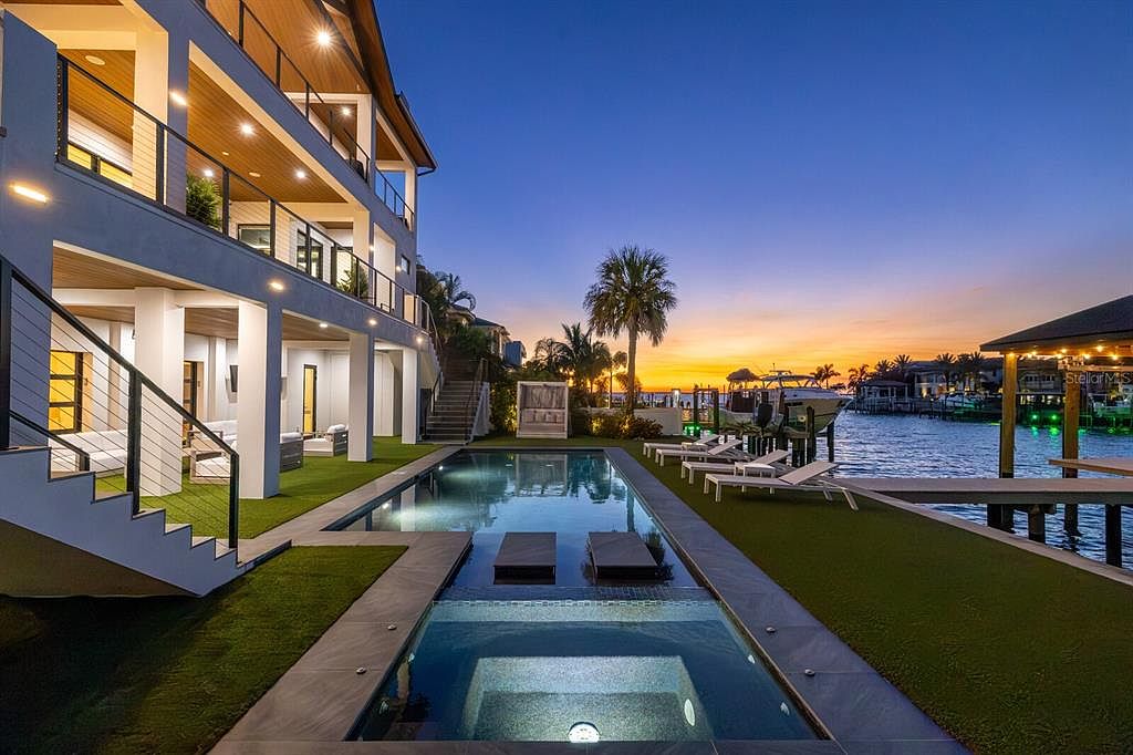 This image showcases a luxurious backyard with a modern pool and spa area, set against a waterfront backdrop at sunset. The multi-level house features balconies and sleek architectural lines, complemented by manicured green spaces and lounge chairs. The scene evokes a sense of high-end living and relaxation.