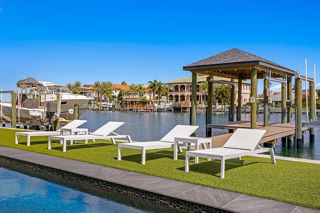 This exterior shot showcases a luxurious waterfront property with a focus on outdoor living. The image features a modern pool with a sleek tile edge, leading to a manicured green space furnished with white lounge chairs. A covered dock with a boat lift and a boat are visible, along with waterfront homes in the background, creating an impression of upscale coastal living.