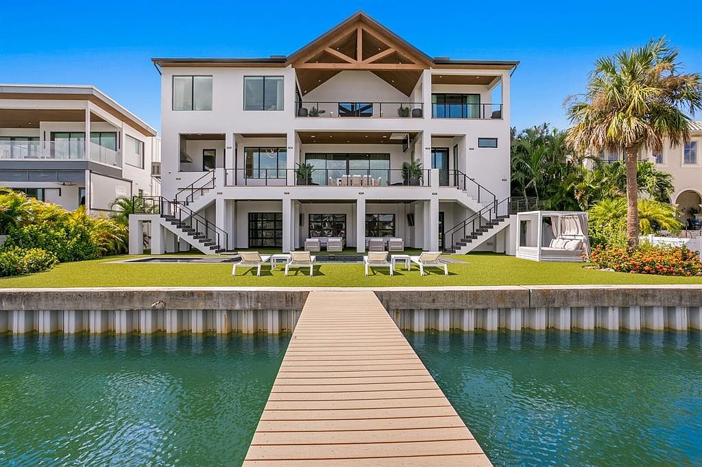 This is a rear exterior view of a modern, multi-story waterfront home. The house features multiple balconies, large windows, and a symmetrical design with staircases leading down to the lawn. A wooden dock extends into the water, and lounge chairs are arranged on the manicured lawn, suggesting a luxurious outdoor living space.