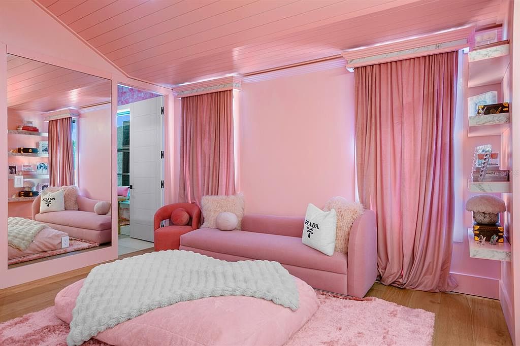 This is a pink-themed bedroom featuring a pink sofa, pink walls, and pink curtains. A large mirror reflects the room, creating an illusion of more space. The room is decorated with plush pillows and a soft rug, giving it a cozy and luxurious feel.