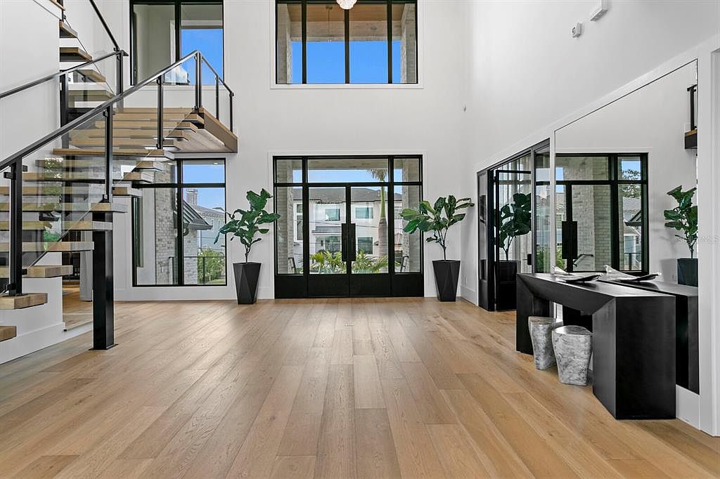 This is an interior shot of a grand entryway featuring a modern staircase with glass railings and wooden steps. The space boasts high ceilings, large windows with black frames, and light wood flooring. Two potted plants flank the front door, adding a touch of greenery to the minimalist design.