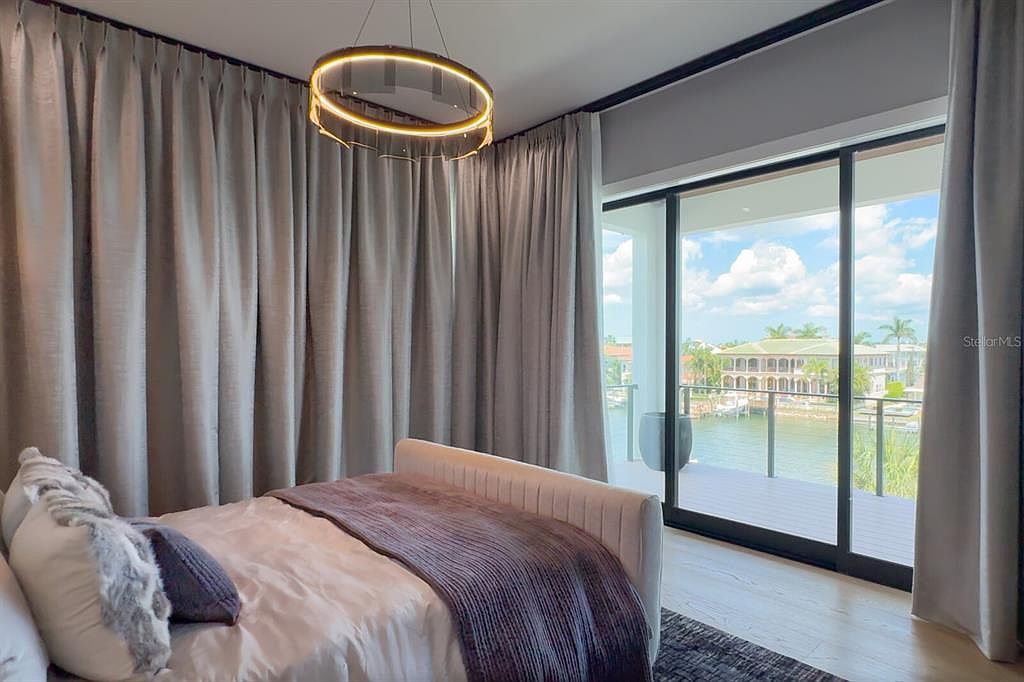 This is a primary bedroom featuring a modern aesthetic with neutral tones. The room includes a bed with decorative pillows and a throw blanket, a contemporary circular light fixture, and floor-to-ceiling curtains that frame a large sliding glass door leading to a balcony with water views. The overall impression is luxurious and serene.