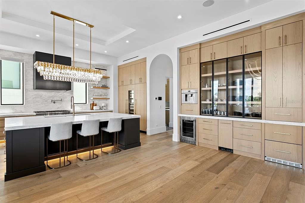 This is a bright and modern kitchen featuring a large island with seating, custom cabinetry, and high-end appliances. The kitchen has a clean and luxurious feel, with a focus on functionality and style. The hardwood floors add warmth to the space, and the open layout is perfect for entertaining.