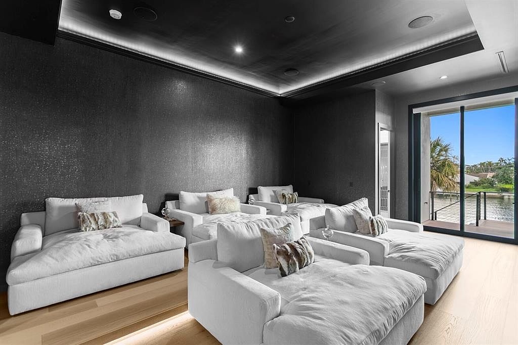 This interior shot showcases a modern living room or home theater setup with multiple white chaise lounges arranged for comfortable viewing. The room features a dark, textured wall and ceiling with recessed lighting, creating a cinematic atmosphere. A large sliding glass door provides natural light and a view of the outdoor scenery, blending indoor and outdoor spaces.