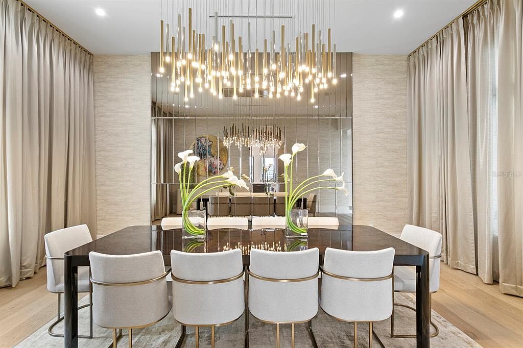 This is an interior shot of a dining room featuring a large, dark-colored dining table surrounded by eight chairs with light-colored upholstery and gold accents. Above the table hangs a modern chandelier with multiple light fixtures, and a mirrored wall behind the table reflects the chandelier and the room, creating a sense of spaciousness. The room is flanked by floor-to-ceiling curtains, adding a touch of elegance.