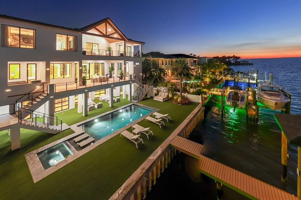 This image showcases the rear exterior of a luxurious waterfront property at dusk. The multi-level home features balconies, a swimming pool, a spa, and a private dock with boats. The manicured lawn, outdoor lounge areas, and strategic lighting create an inviting and upscale ambiance.