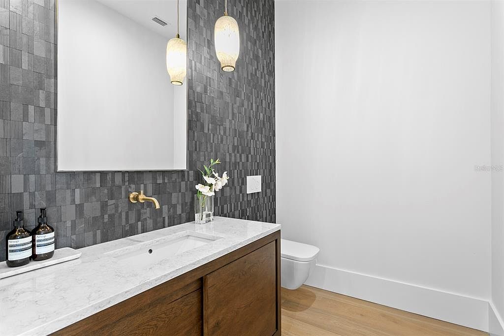 This is a modern guest bathroom featuring a sleek vanity with a wood-grain finish and a white countertop. A rectangular mirror hangs above the sink, complemented by stylish pendant lights. The wall behind the vanity is covered in a textured gray tile, while the adjacent wall is painted white, creating a clean and contemporary aesthetic.