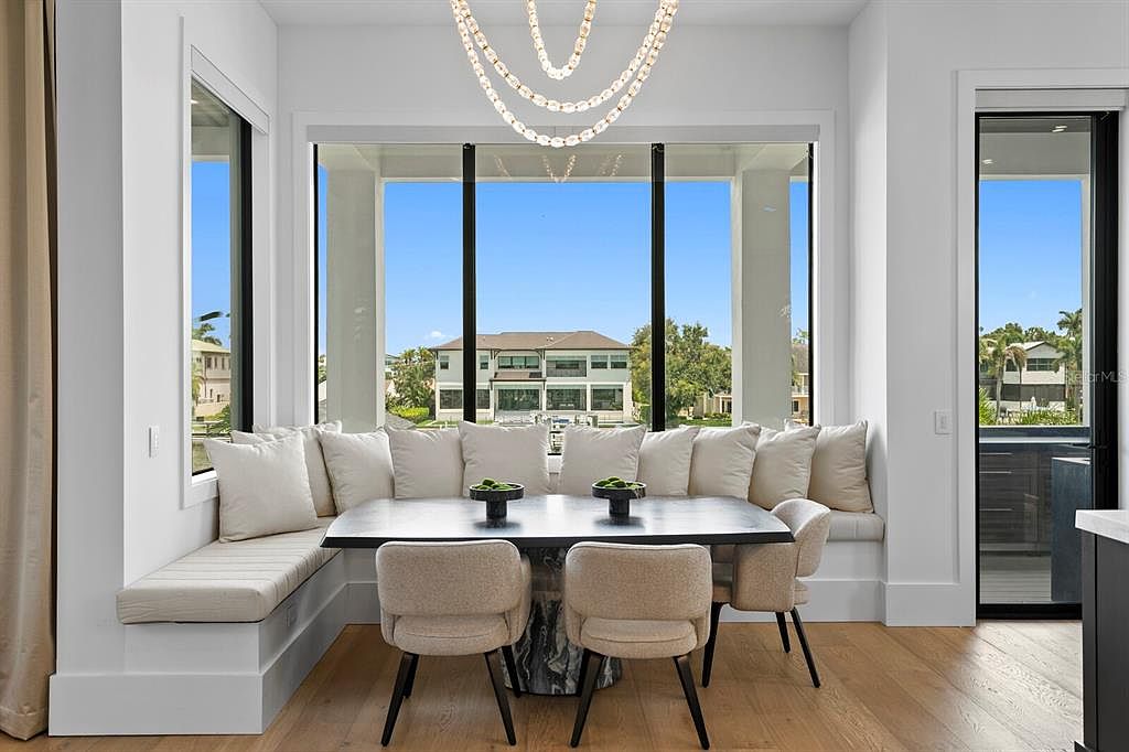 This is an interior shot of a modern dining room featuring a built-in bench seat with cushions and pillows, a dark-topped table with a unique base, and four upholstered chairs. Large windows offer a view of a waterfront property, and a decorative chandelier hangs above the table. The room has a bright and airy feel with hardwood floors.