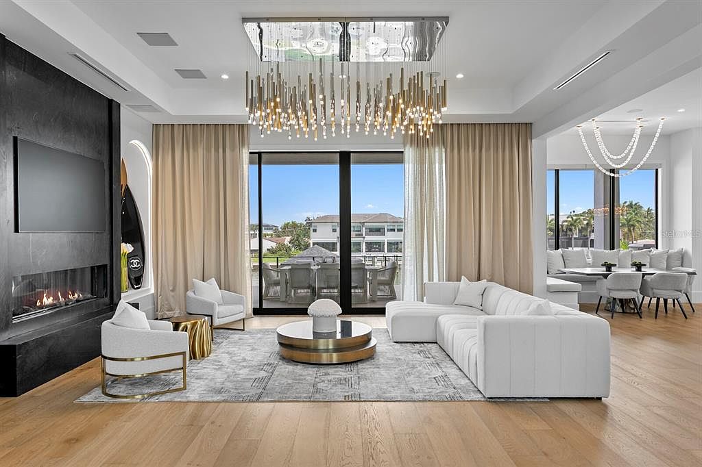 This is a luxurious living room featuring a modern fireplace with a large television above, a white sectional sofa, and two armchairs arranged around a round coffee table on a patterned rug. Large windows with beige curtains offer a view of the outdoors, and a striking chandelier hangs from the ceiling. The perspective is a wide shot, showcasing the room's spaciousness and elegant design.