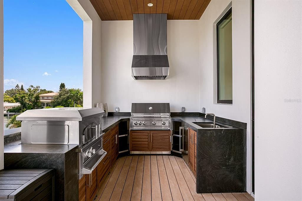 This image showcases an outdoor kitchen area on a patio or balcony. The space features a built-in grill, stainless steel appliances, dark countertops, and wooden cabinetry, creating a modern and functional outdoor cooking and entertaining area. The wooden flooring adds warmth, while the open view provides a pleasant backdrop.