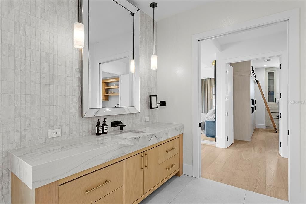 This is a well-lit primary bathroom featuring a modern vanity with a marble countertop and light wood cabinetry. The wall is adorned with textured tiles, and the space is illuminated by pendant lights. An open doorway leads to a bedroom and closet area, creating a sense of flow and spaciousness.