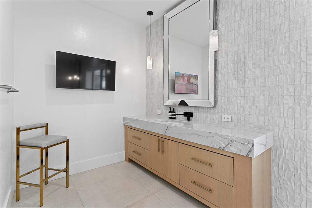 This is a modern guest bathroom featuring a light wood vanity with a white marble countertop and gold hardware. A large mirror hangs above the vanity, reflecting a television mounted on the wall. The walls are a mix of textured white tile and smooth white paint, complemented by a gold-framed stool, creating a clean and stylish aesthetic.