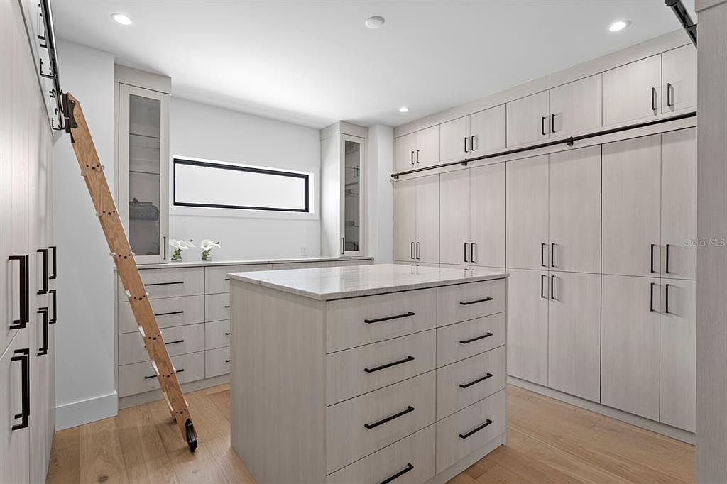 This is a spacious walk-in closet featuring custom cabinetry with a light wood finish and sleek black hardware. A central island provides additional storage and countertop space, while a rolling ladder offers access to higher shelves. The closet is well-lit and organized, creating a luxurious and functional space.