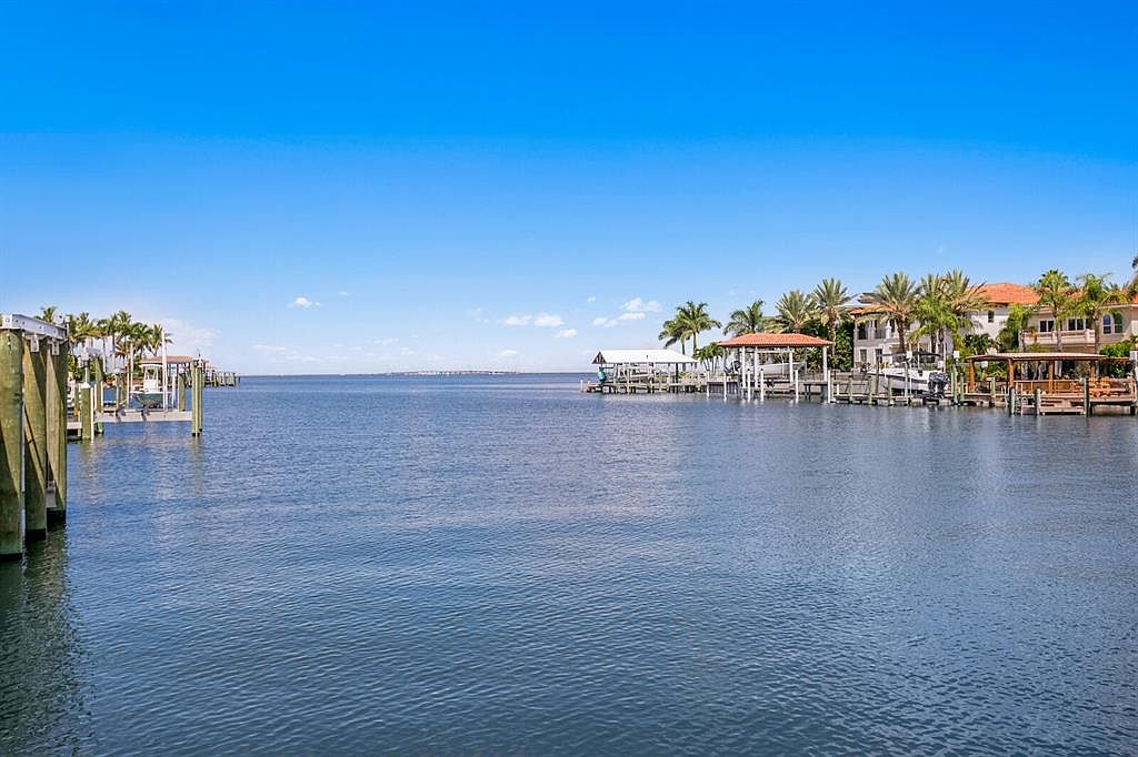 This exterior shot showcases the rear of a waterfront property, featuring multiple docks and boat lifts. The architecture includes Mediterranean-style homes with terracotta roofs and lush palm trees, creating a luxurious coastal ambiance. The calm water and clear blue sky enhance the appeal of this waterfront estate.