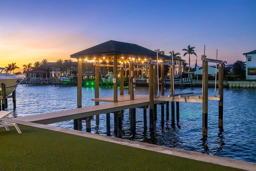 This image showcases the rear exterior of a waterfront property at dusk, featuring a private dock with a covered structure illuminated by string lights. The dock extends into the water, providing boat access and a relaxing space. The property is surrounded by lush landscaping and other upscale homes, creating a luxurious and serene atmosphere.