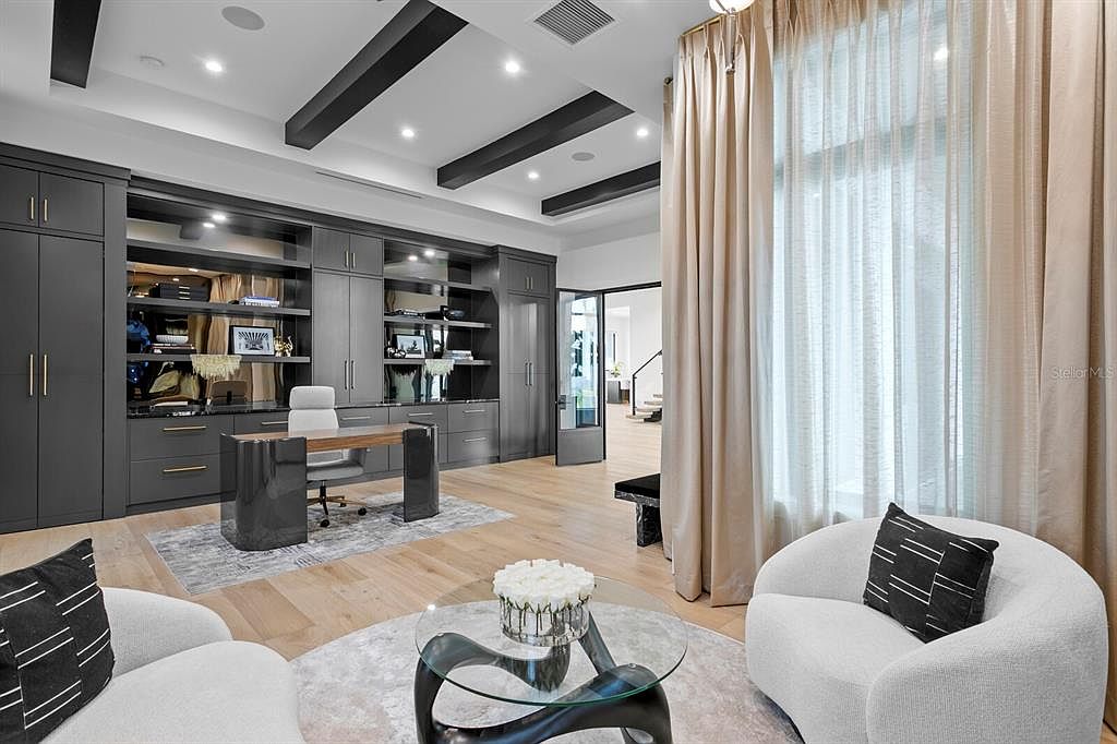 This is an interior shot of a modern office space featuring custom built-in cabinetry in a dark gray finish, providing ample storage and display areas. A sleek desk and comfortable seating arrangement create an inviting workspace, complemented by a stylish rug and a glass coffee table with floral arrangement. The room is well-lit with natural light filtering through sheer curtains, enhancing the sophisticated and functional design.
