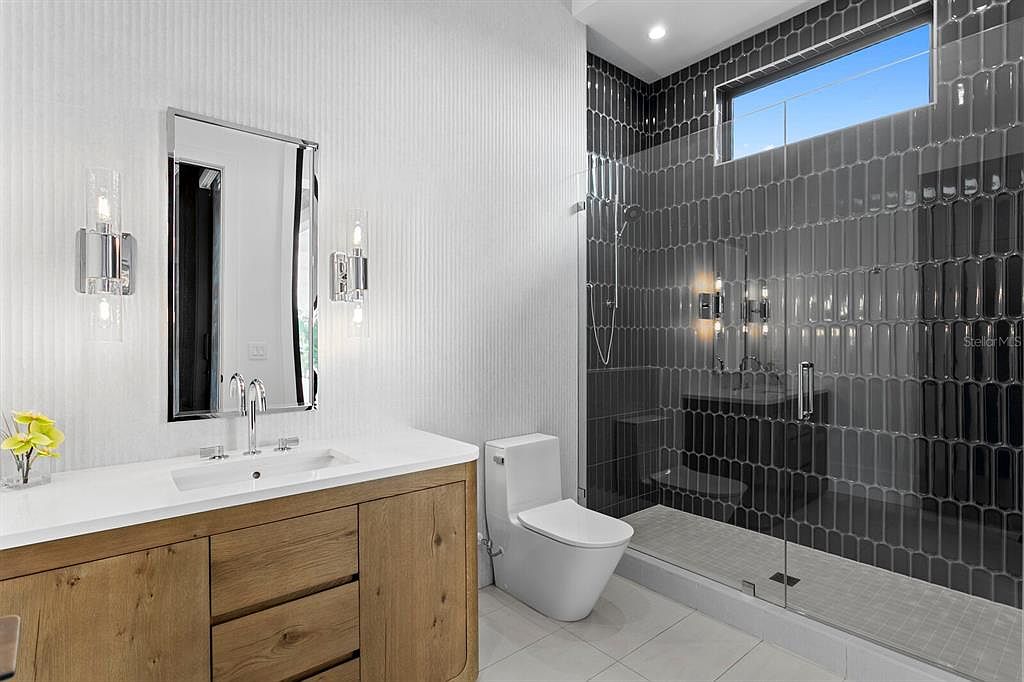 This is a modern primary bathroom featuring a wood-grain vanity with a white countertop and a rectangular sink. A large mirror hangs above the sink, flanked by sconce lighting. The shower is enclosed with glass and features dark, vertically stacked tiles, creating a sleek and contemporary look. A toilet is positioned next to the vanity.