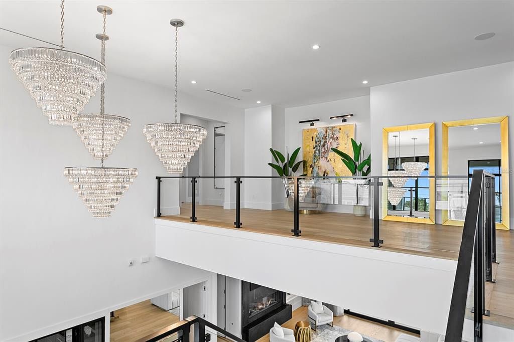 This interior shot showcases a luxurious hallway and staircase area, featuring modern design elements. The space is illuminated by several elaborate crystal chandeliers, adding a touch of elegance. A glass-paneled railing provides a clear view of the lower level, where a fireplace and seating area are visible, creating an open and inviting atmosphere.