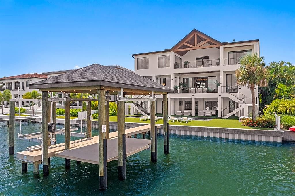 This is a rear view of a modern, multi-story waterfront home. The house features multiple balconies, large windows, and a staircase leading down to a well-manicured lawn. A private dock with a covered boat lift extends into the water, enhancing the property's appeal for boating enthusiasts.